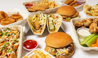 A variety of takeout food in white containers, including burgers, tacos, fries, and chicken wings.