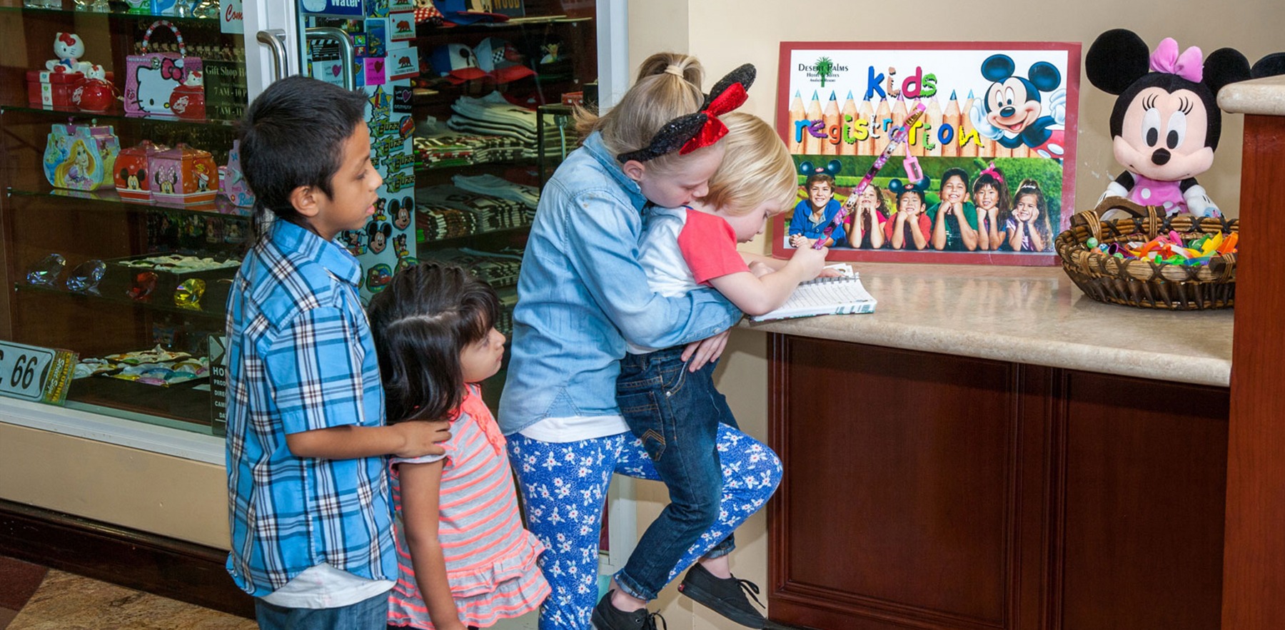 A family of four, including two children, stand at a counter with a Minnie Mouse stuffed animal and a poster that reads 'kids registration'.